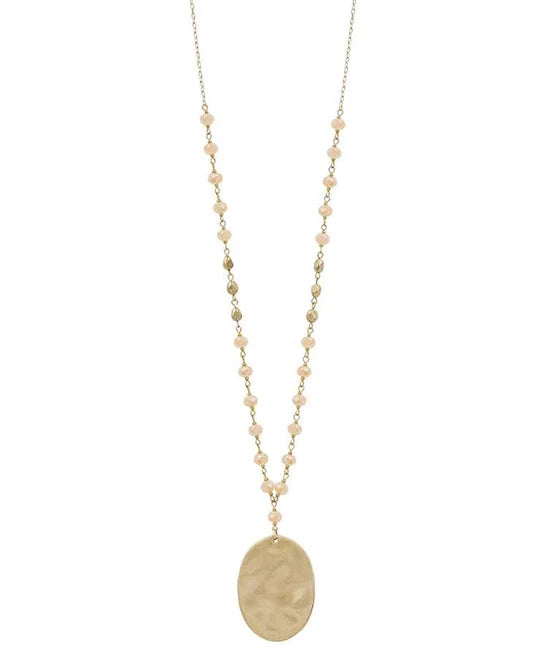 A long gold-tone chain necklace featuring small peach-colored beads spaced along the chain and a large, flat oval pendant with a textured finish, shown against a plain white background.