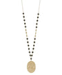 A long beaded necklace featuring a hammered oval gold pendant, accented by small black and gold beads on a delicate chain, shown against a plain white background.
