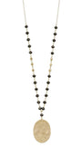 A long beaded necklace featuring a hammered oval gold pendant, accented by small black and gold beads on a delicate chain, shown against a plain white background.