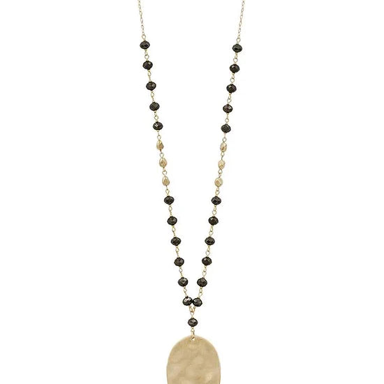 A long beaded necklace featuring a hammered oval gold pendant, accented by small black and gold beads on a delicate chain, shown against a plain white background.