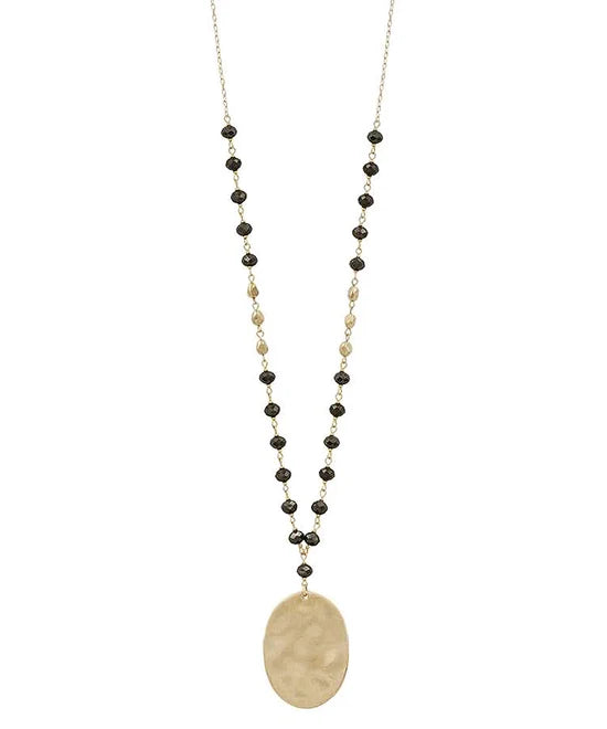 A long beaded necklace featuring a hammered oval gold pendant, accented by small black and gold beads on a delicate chain, shown against a plain white background.