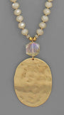 A gold-tone pendant necklace featuring a large hammered oval pendant, a faceted iridescent clear bead, and a beaded chain of cream-colored faceted beads, shown against a solid gray background.