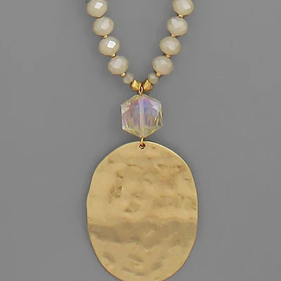 A gold-tone pendant necklace featuring a large hammered oval pendant, a faceted iridescent clear bead, and a beaded chain of cream-colored faceted beads, shown against a solid gray background.