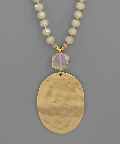 A gold-tone pendant necklace featuring a large hammered oval pendant, a faceted iridescent clear bead, and a beaded chain of cream-colored faceted beads, shown against a solid gray background.