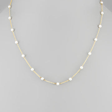 A delicate gold chain necklace featuring evenly spaced, small white pearl beads, displayed on a neutral gray background.