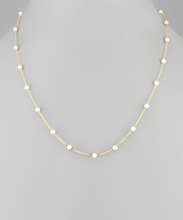A delicate gold chain necklace featuring evenly spaced, small white pearl beads, displayed on a neutral gray background.