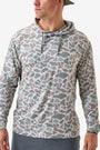 A man wearing a long-sleeve hooded shirt with an all-over abstract camouflage pattern in muted gray, taupe, and beige tones, paired with gray pants against a clean light gray studio background.