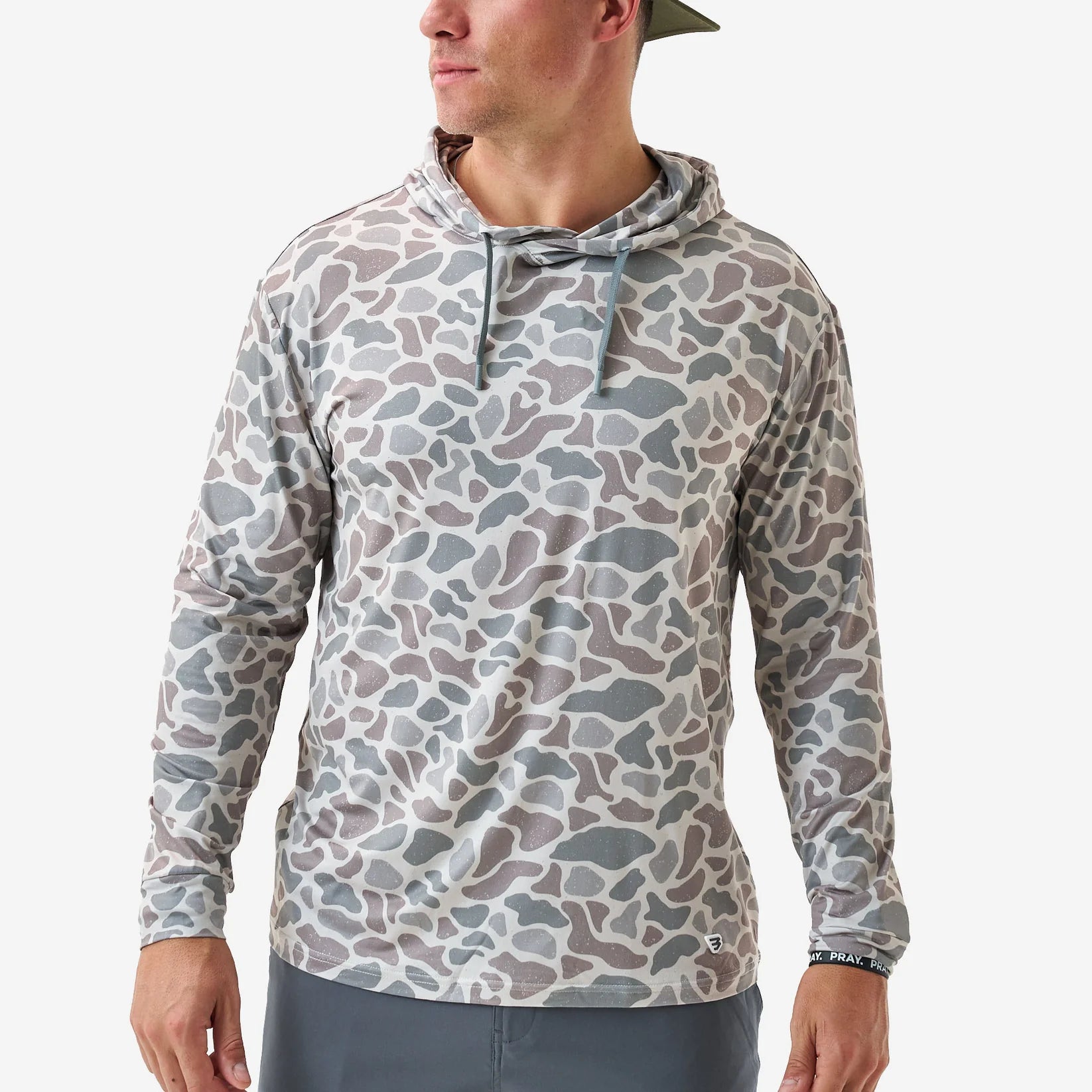 A man wearing a long-sleeve hooded shirt with an all-over abstract camouflage pattern in muted gray, taupe, and beige tones, paired with gray pants against a clean light gray studio background.