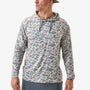 A man wearing a long-sleeve hooded shirt with an all-over abstract camouflage pattern in muted gray, taupe, and beige tones, paired with gray pants against a clean light gray studio background.