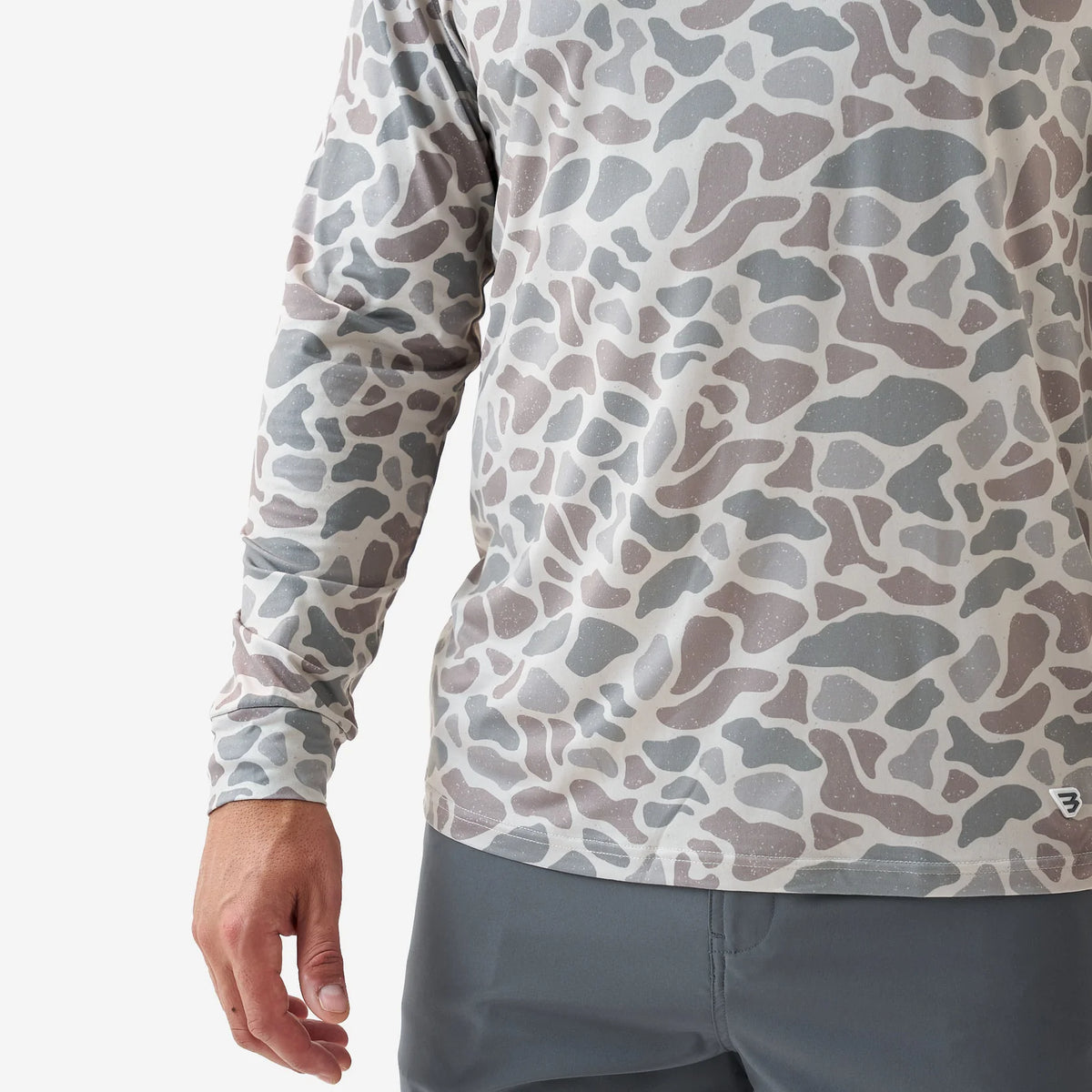 A close-up of a person wearing a long-sleeve performance shirt with an abstract organic camouflage pattern in gray, taupe, and off-white, paired with gray athletic pants.