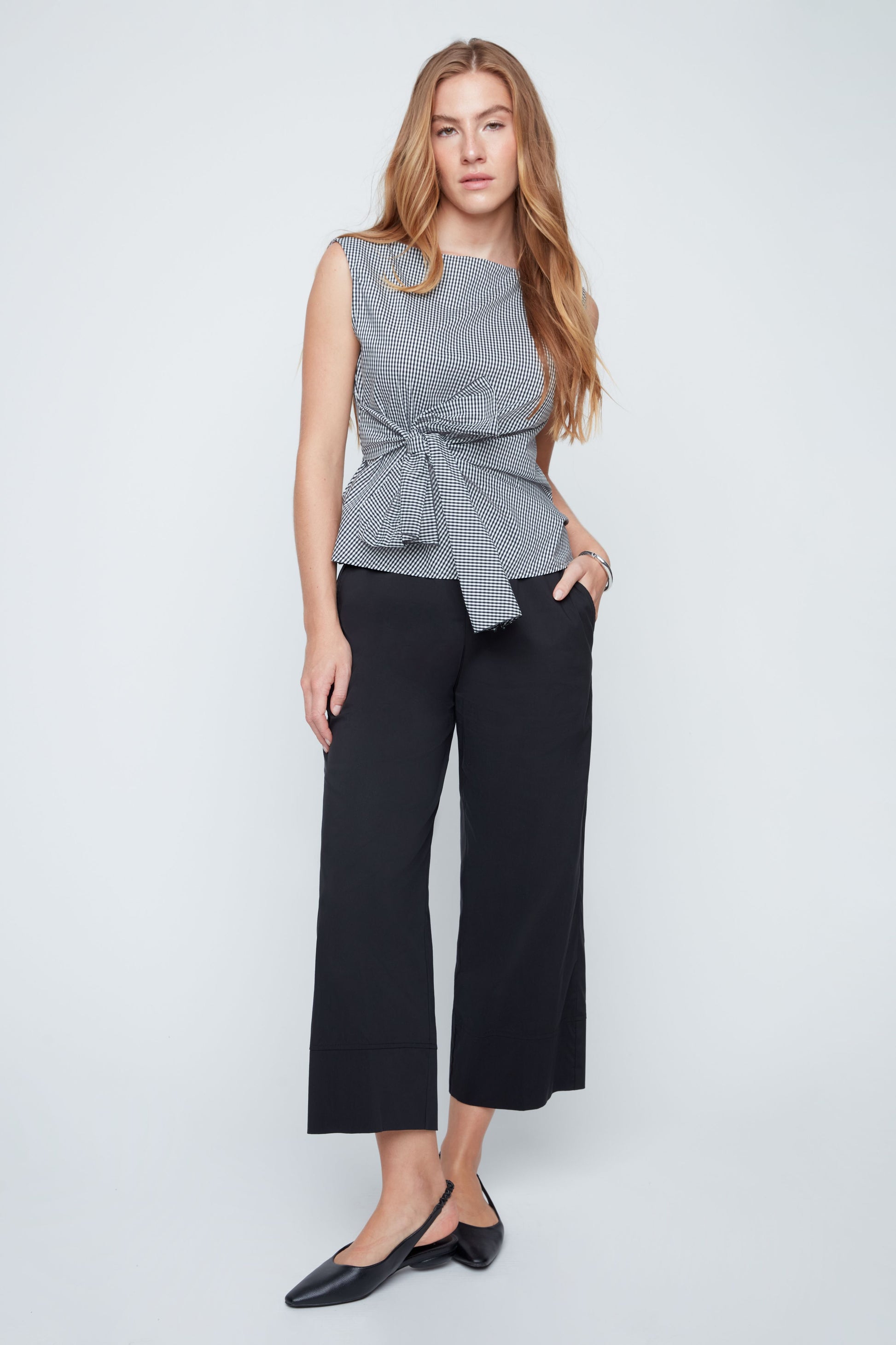 Woman wearing a gray sleeveless top with a knot detail and black pants on a light gray background