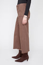 A model shown from the waist down wears brown wide-leg cropped jeans with a black long-sleeve top and dark brown pointed-toe ankle boots, standing against a light gray studio background.