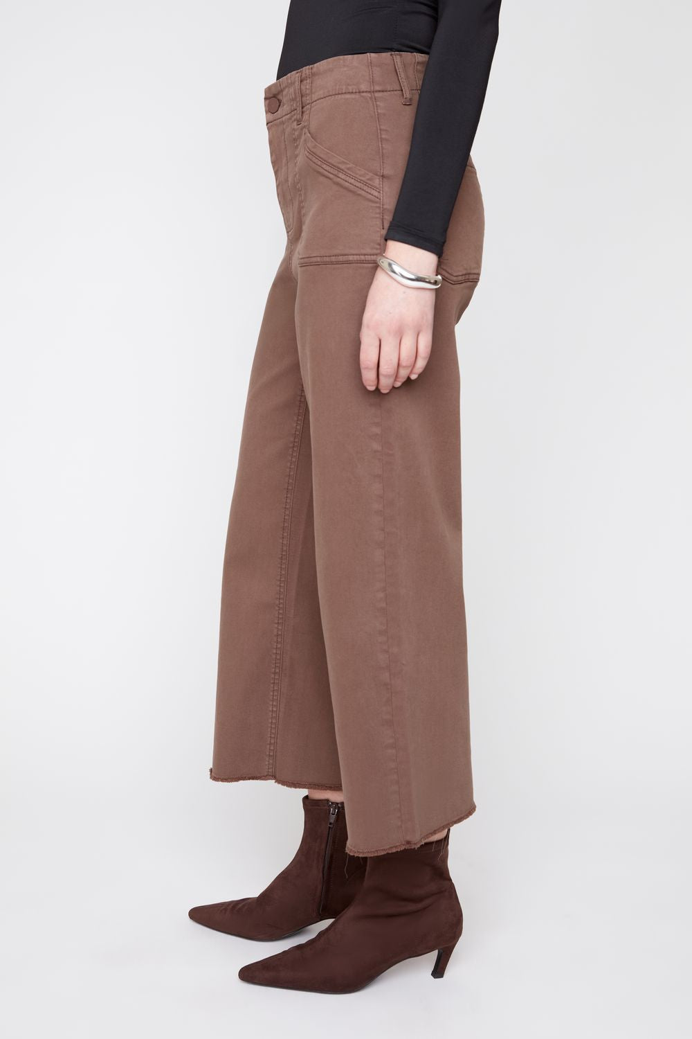 A model shown from the waist down wears brown wide-leg cropped jeans with a black long-sleeve top and dark brown pointed-toe ankle boots, standing against a light gray studio background.