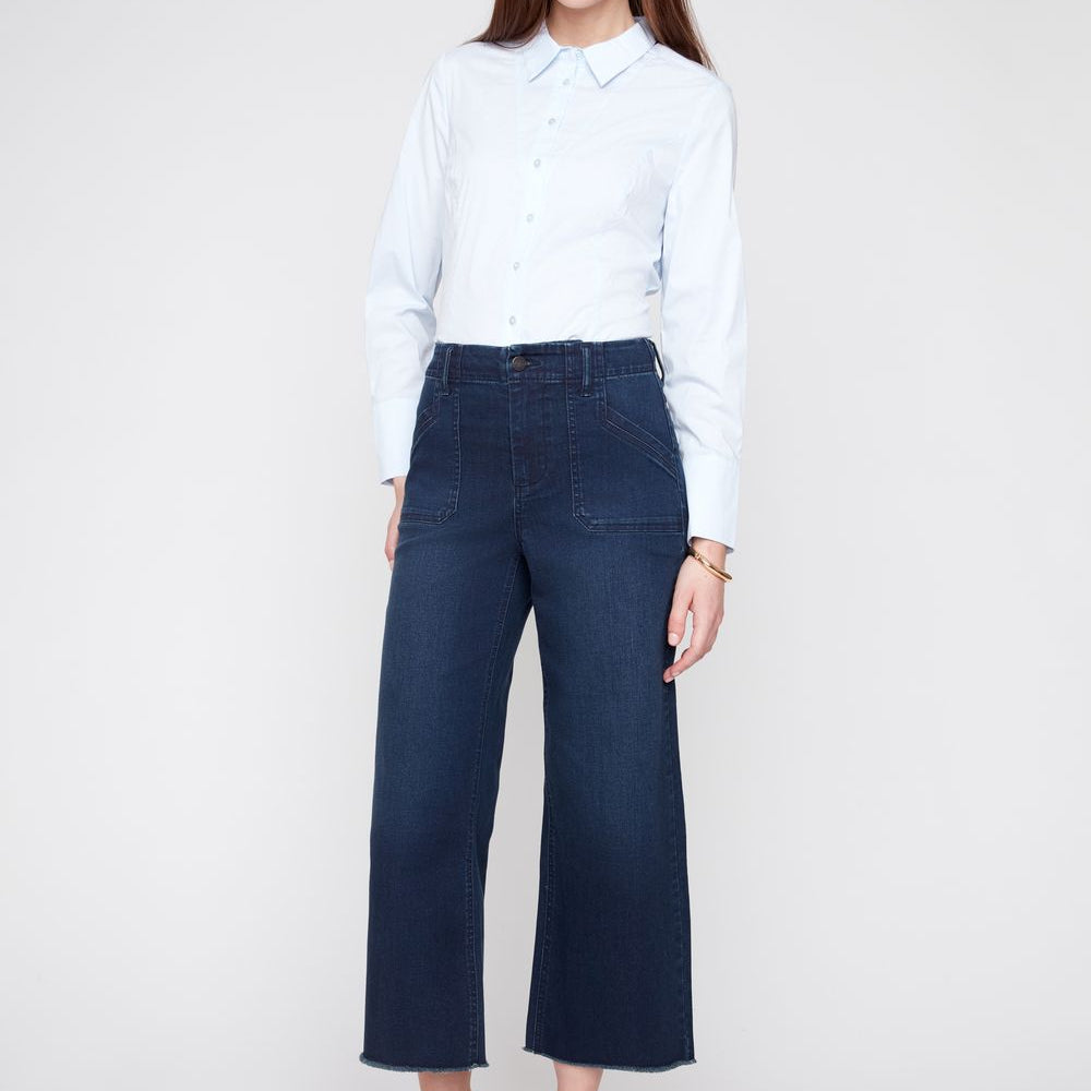 A woman modeling a light blue long-sleeve button-up shirt and dark blue wide-leg cropped jeans, paired with dark red pointed-toe heels, against a light gray studio background.