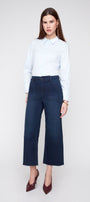 A woman modeling a light blue long-sleeve button-up shirt and dark blue wide-leg cropped jeans, paired with dark red pointed-toe heels, against a light gray studio background.