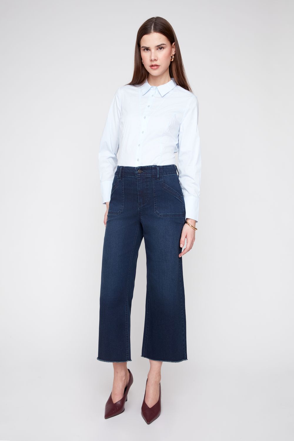 A woman modeling a light blue long-sleeve button-up shirt and dark blue wide-leg cropped jeans, paired with dark red pointed-toe heels, against a light gray studio background.