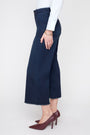 A model shown from the waist down wearing dark navy blue cropped wide-leg denim jeans, a white long-sleeve shirt, and dark burgundy pointed-toe heels against a plain white studio background.