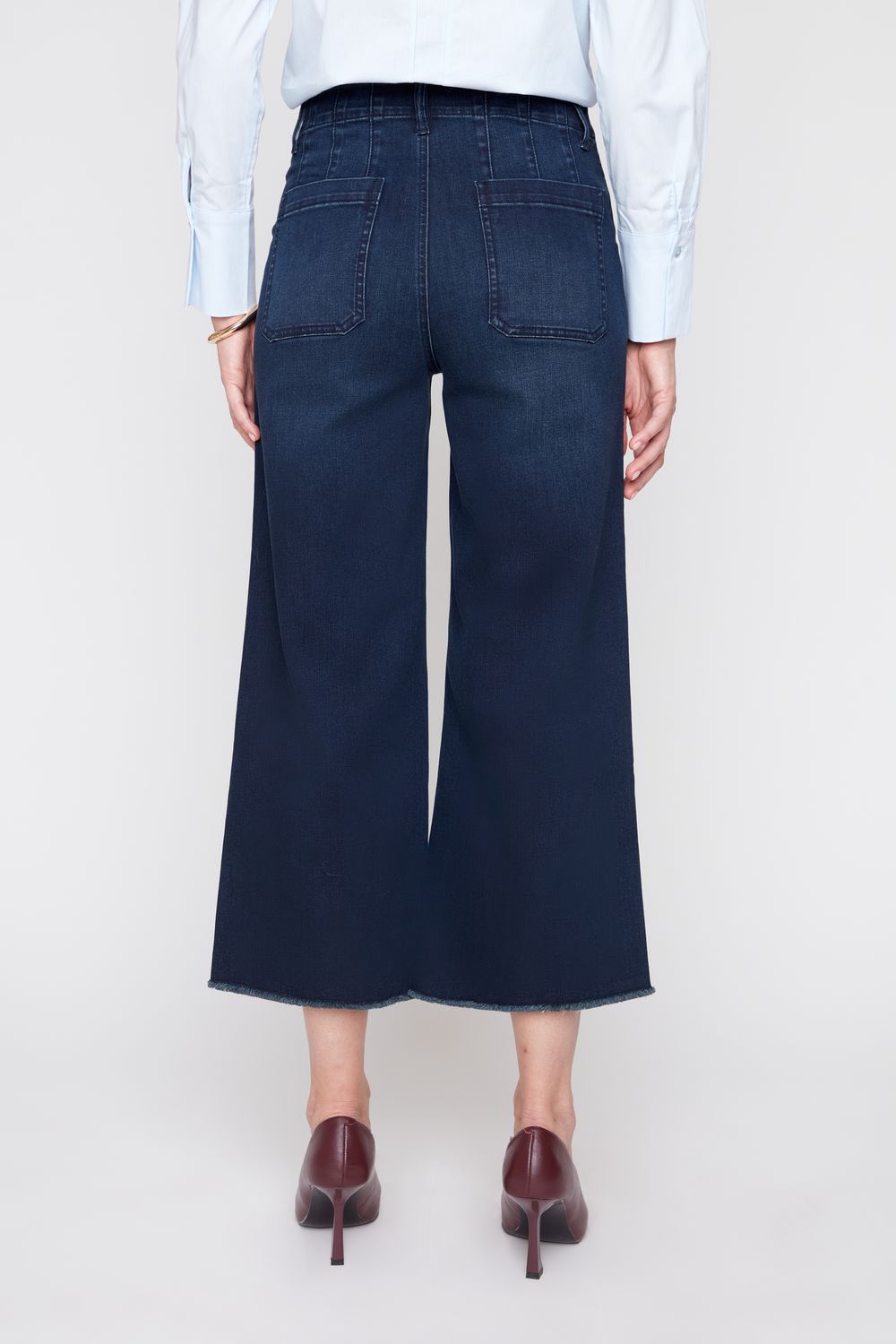 A model shown from the back wearing dark blue wide-leg cropped jeans, a light blue long-sleeve button-up shirt, and dark brown pointed-toe heels against a plain light gray studio background.