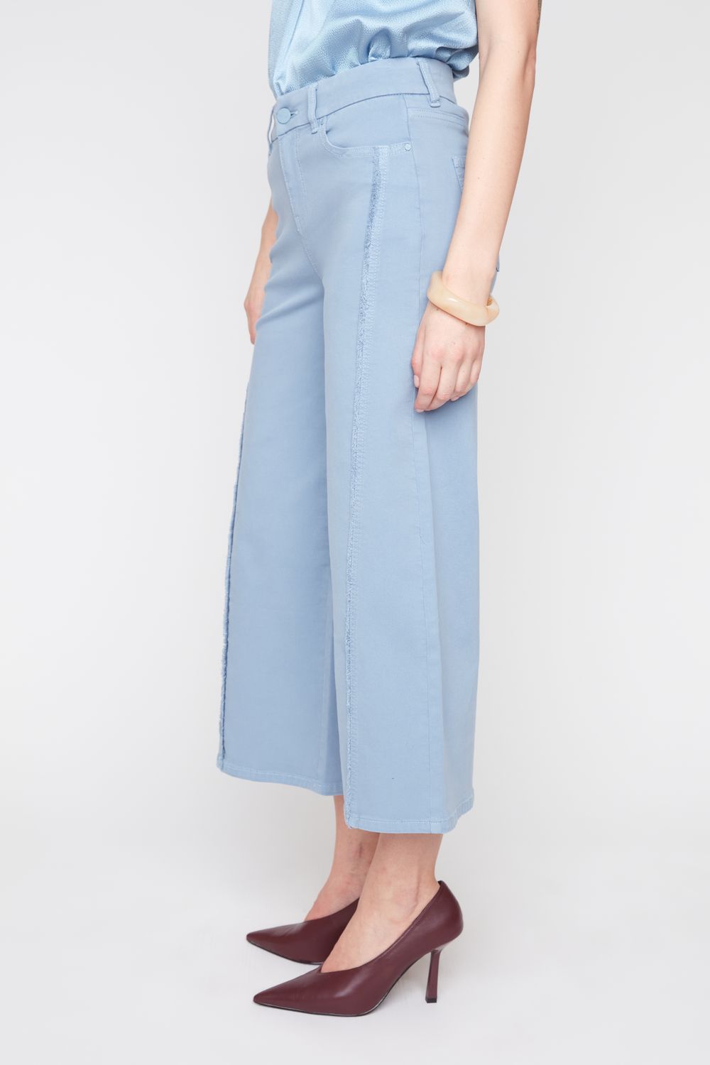 A model wearing light blue wide-leg cropped jeans, a light blue satin top, and dark burgundy pointed-toe heels, shown from the waist down against a plain light gray studio background.
