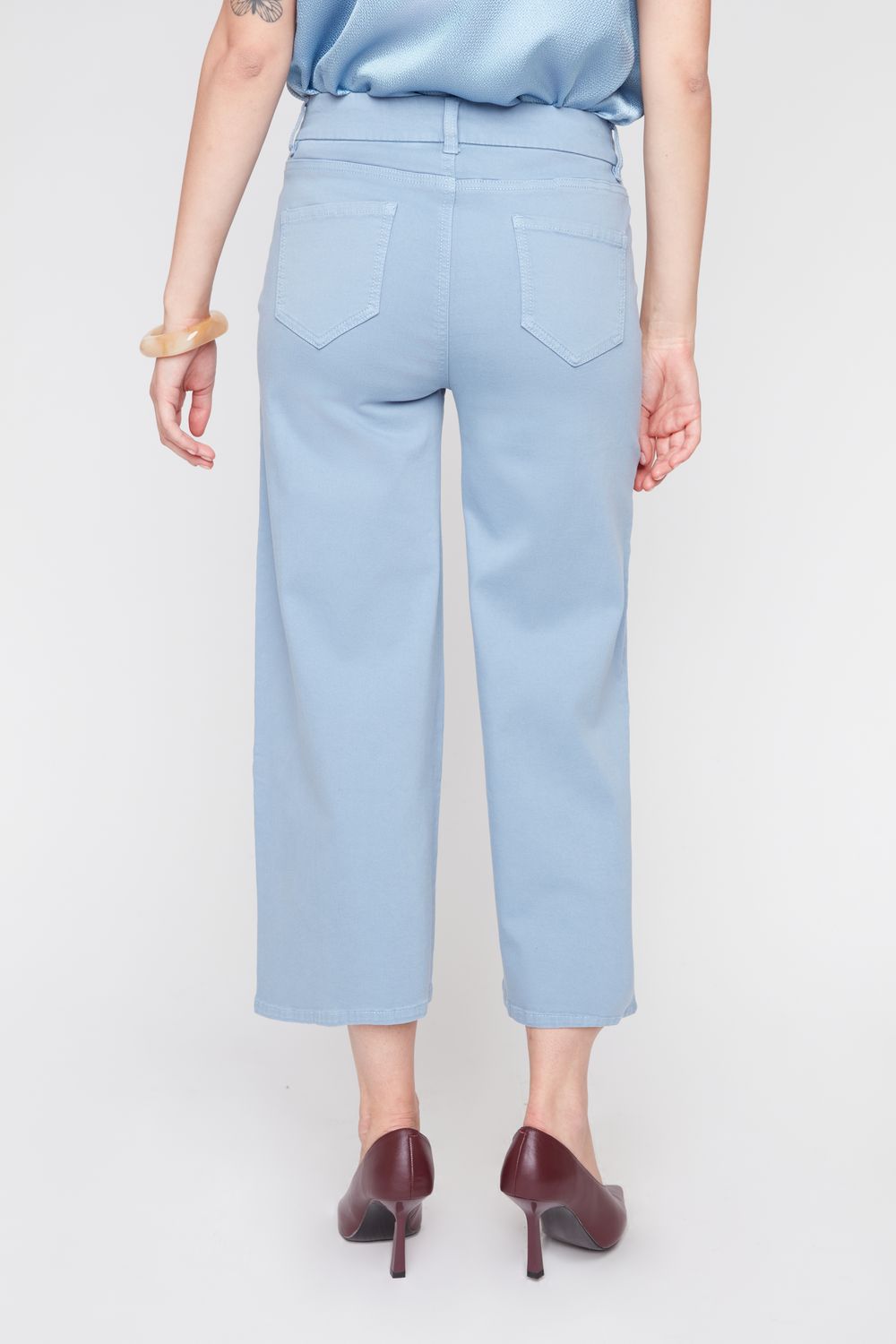A model shown from the back wearing light blue cropped trousers, a light blue satin top, and dark burgundy heels against a plain light gray studio background.