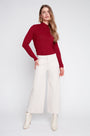 A woman modeling a long-sleeve ribbed knit mock neck top in deep red, paired with off-white wide-leg cropped pants and white ankle boots, against a light gray studio background.