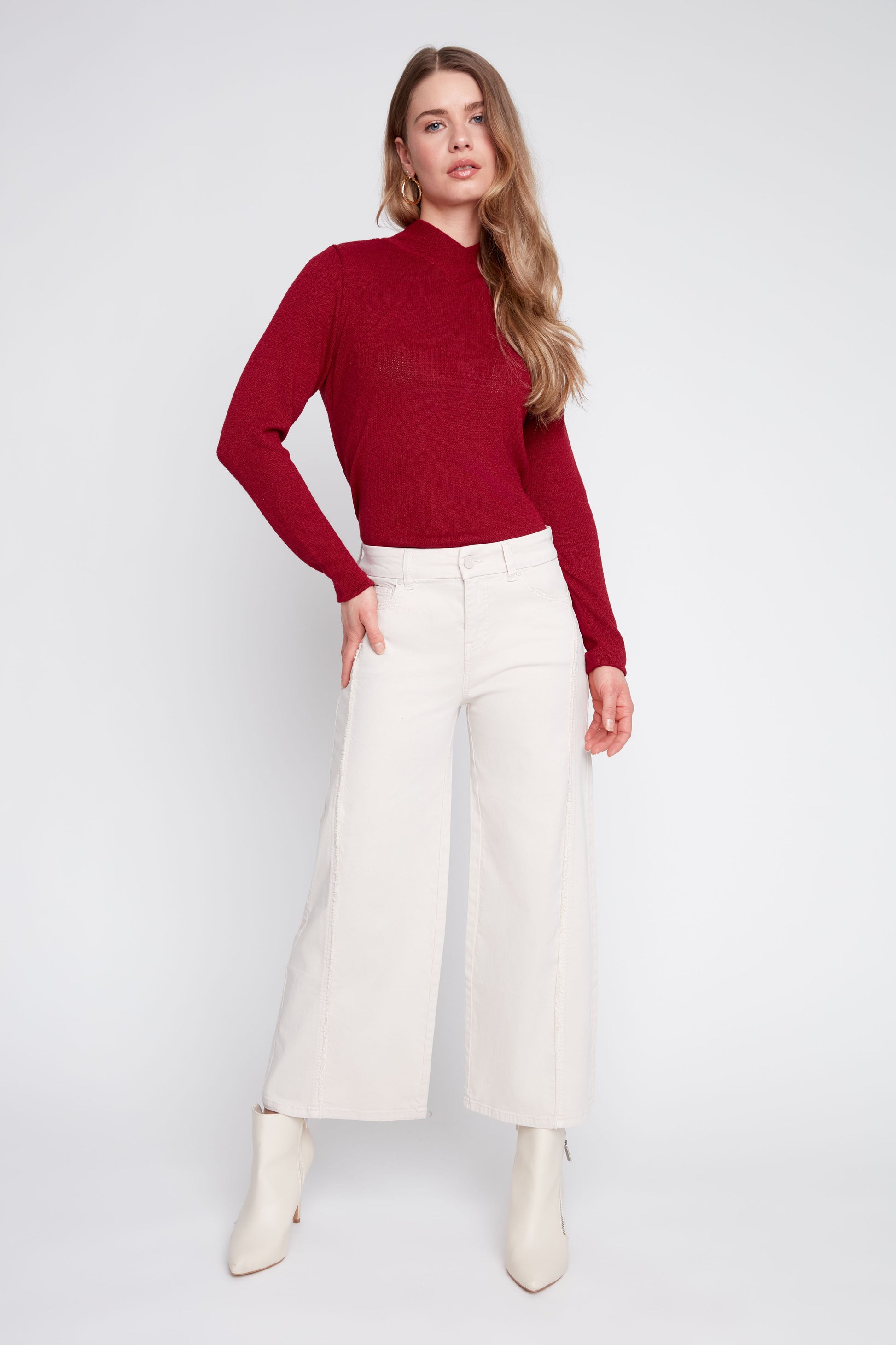 A woman modeling a long-sleeve ribbed knit mock neck top in deep red, paired with off-white wide-leg cropped pants and white ankle boots, against a light gray studio background.