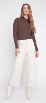 A woman modeling a brown mock-neck long-sleeve top paired with off-white wide-leg trousers and white ankle boots, shot against a light gray studio background.