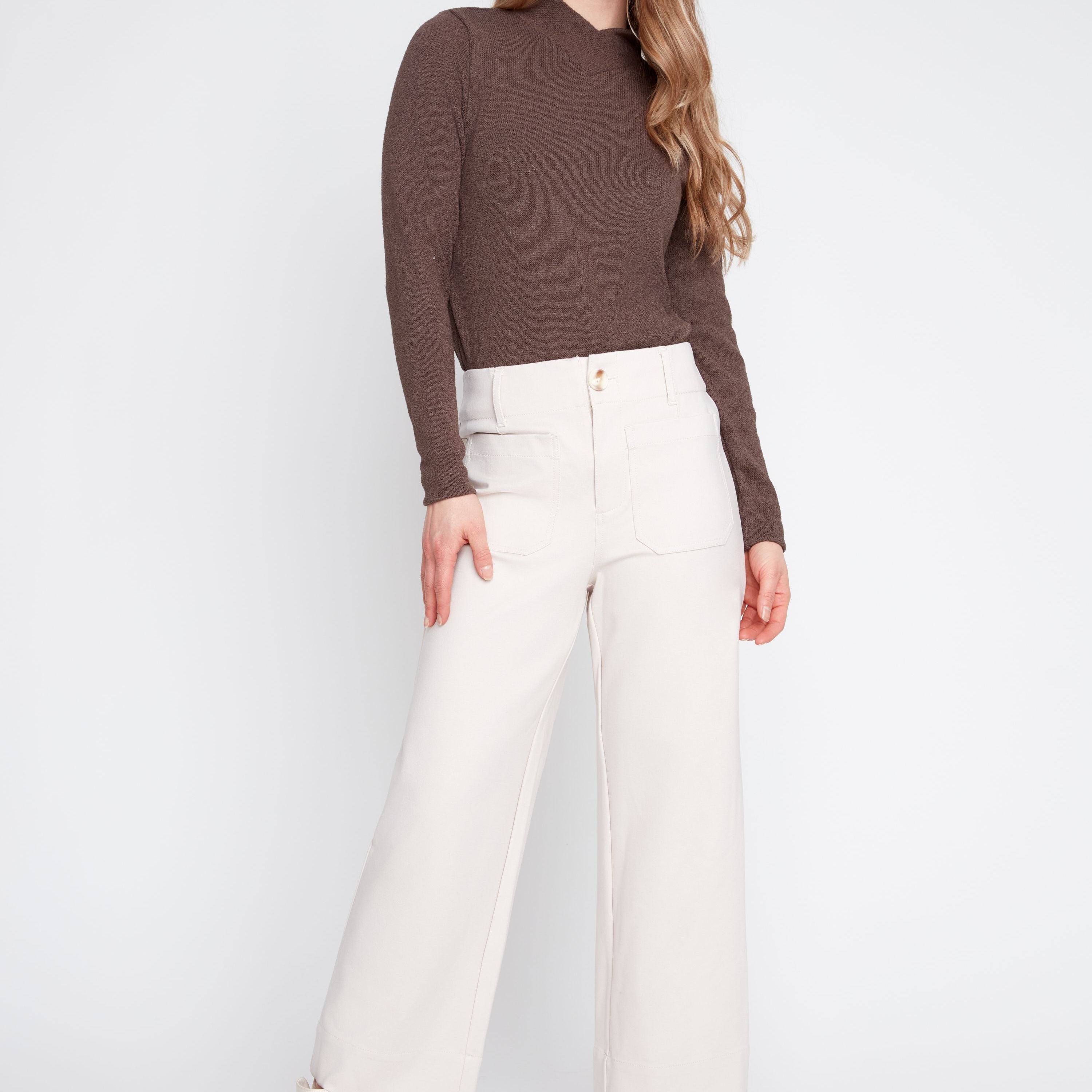 A woman modeling a brown mock-neck long-sleeve top paired with off-white wide-leg trousers and white ankle boots, shot against a light gray studio background.