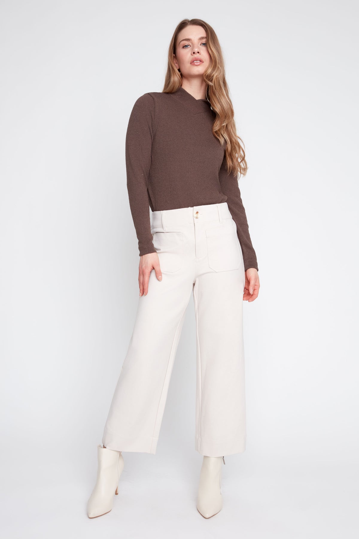 A woman modeling a brown mock-neck long-sleeve top paired with off-white wide-leg trousers and white ankle boots, shot against a light gray studio background.