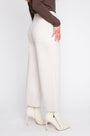 A model is shown from the side wearing off-white wide-leg trousers, a brown long-sleeve top, and off-white pointed-toe ankle boots with a side zipper, set against a plain light gray studio background.