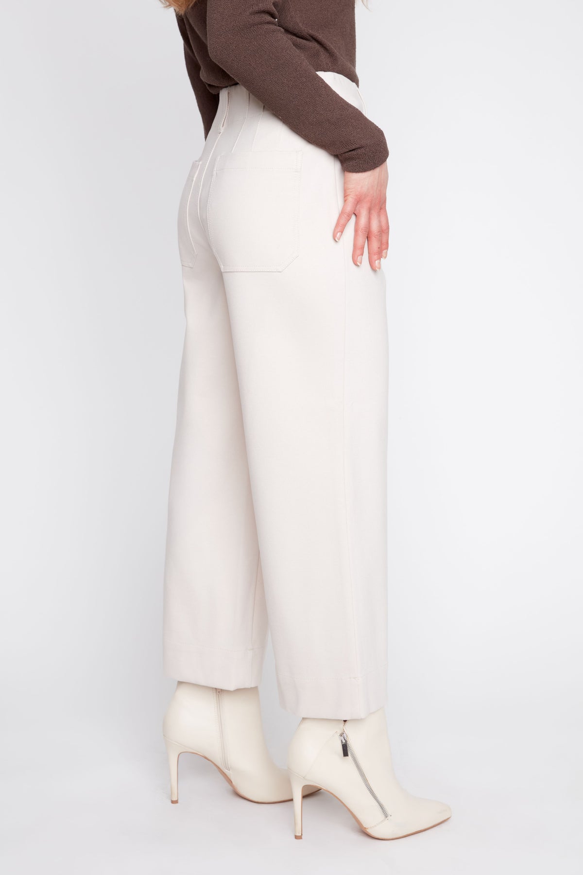 A model is shown from the side wearing off-white wide-leg trousers, a brown long-sleeve top, and off-white pointed-toe ankle boots with a side zipper, set against a plain light gray studio background.