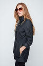 Woman wearing a black raincoat and sunglasses on a light gray background