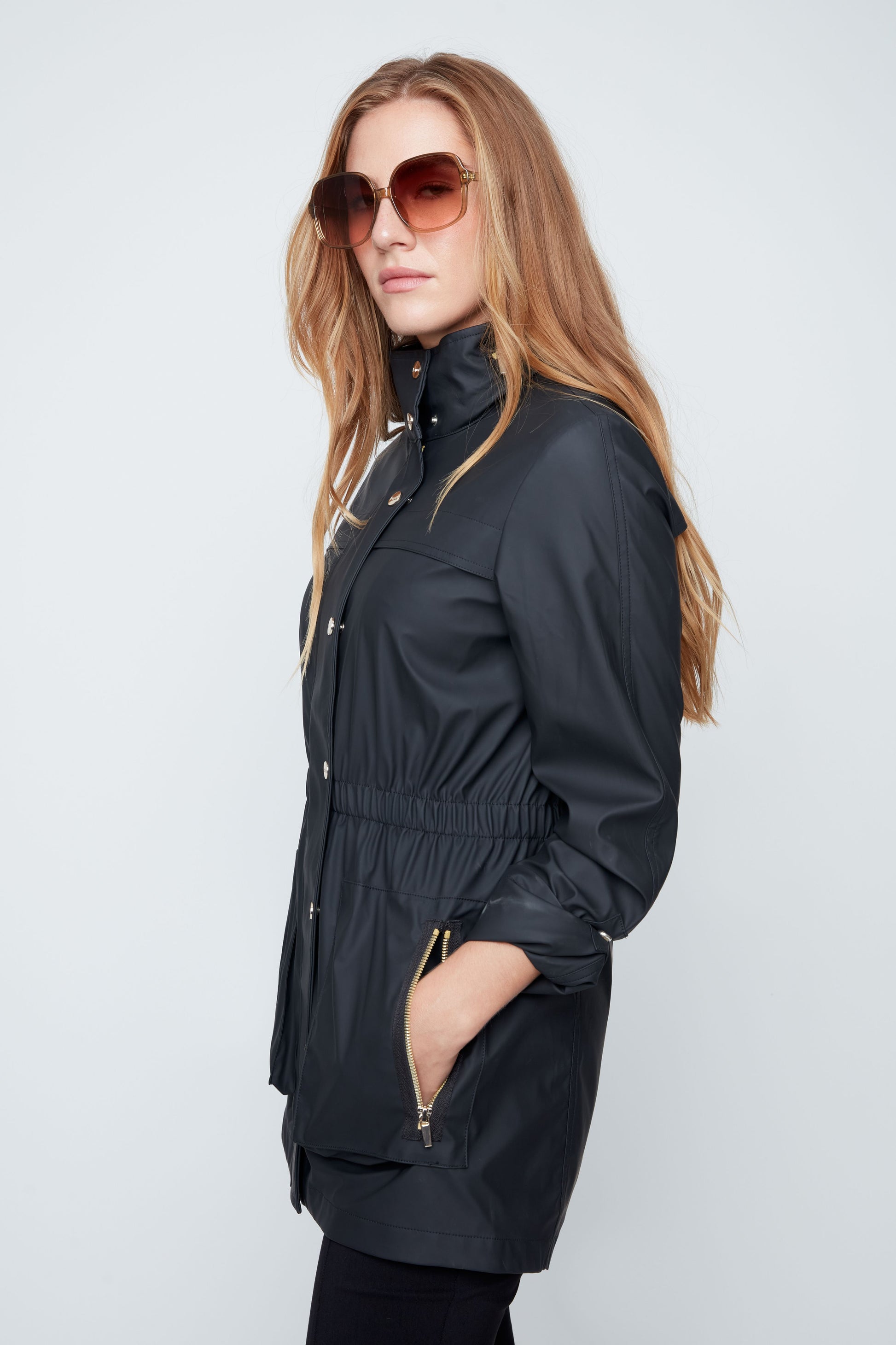 Woman wearing a black raincoat and sunglasses on a light gray background