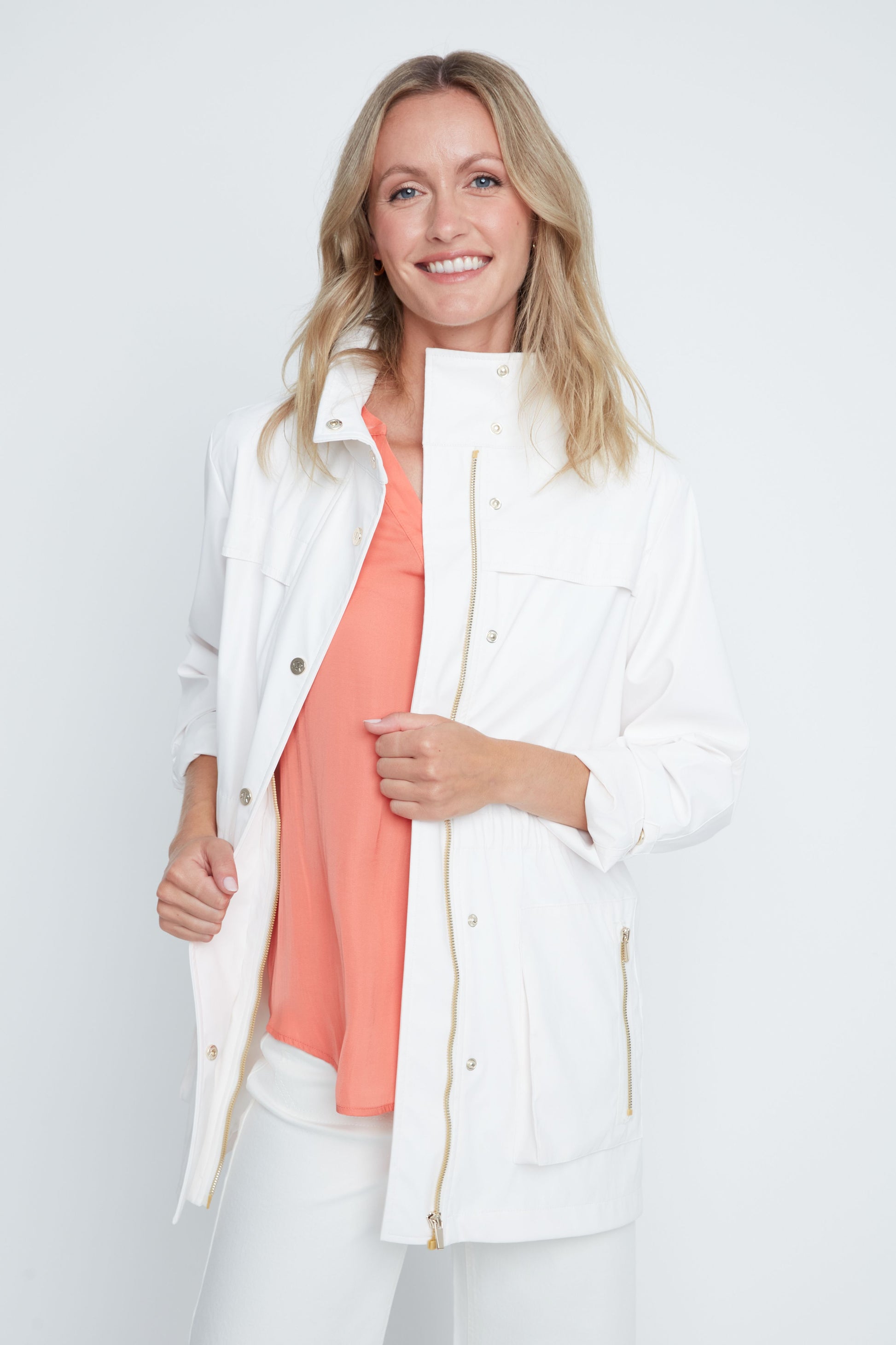 Woman wearing a white jacket over a coral top and white pants on a light gray background
