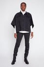 A model stands against a light gray studio background wearing a black cropped denim cape jacket over a white turtleneck, black pants, and black over-the-knee boots.
