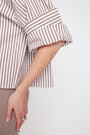 A close-up of a person wearing a brown and beige vertically striped button-up shirt with rolled-up sleeves, paired with beige trousers, against a plain light background.