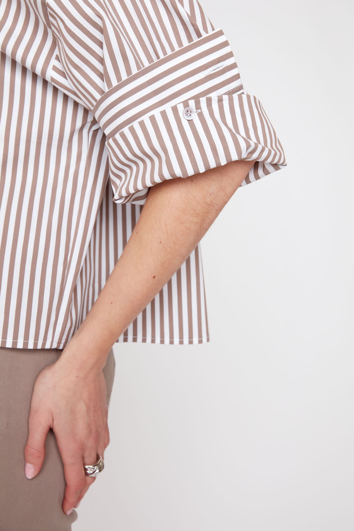 A close-up of a person wearing a brown and beige vertically striped button-up shirt with rolled-up sleeves, paired with beige trousers, against a plain light background.