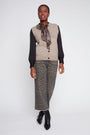 A model stands against a light gray studio background wearing a beige knit vest over a black long-sleeve top, a patterned scarf, wide-leg cropped trousers with a brown and gray animal print, and black ankle boots.