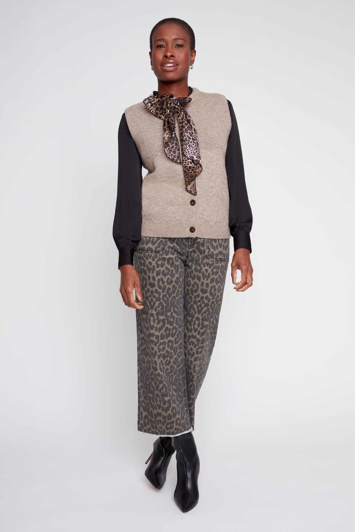 A model stands against a light gray studio background wearing a beige knit vest over a black long-sleeve top, a patterned scarf, wide-leg cropped trousers with a brown and gray animal print, and black ankle boots.