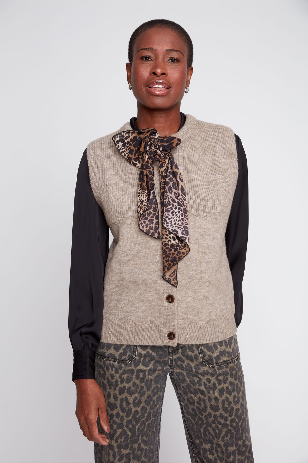 Woman wearing a beige vest over a black long-sleeve shirt with a leopard print scarf and pants on a white background