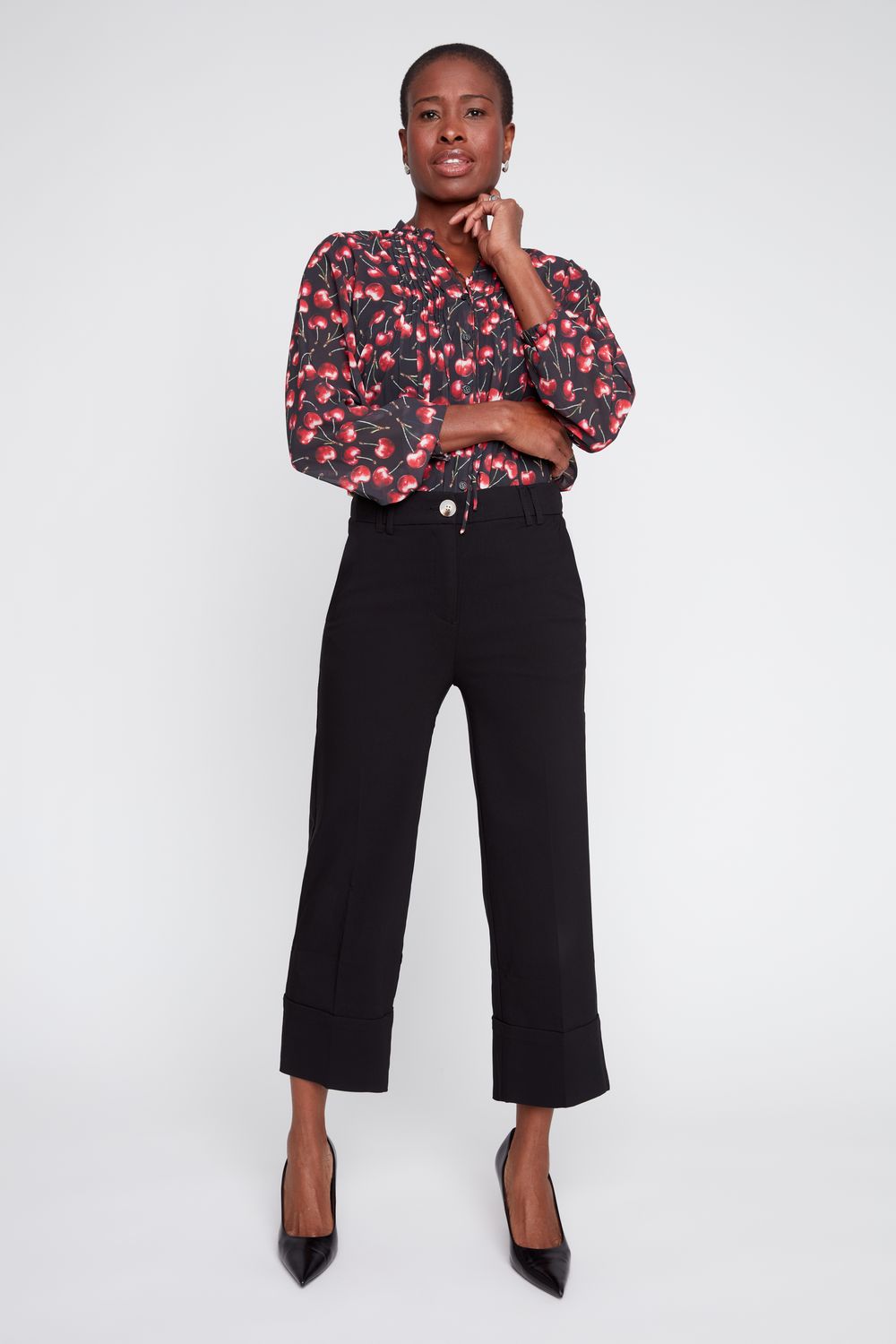 A model stands against a light gray studio background wearing a black cropped wide-leg trouser with a button waist and a red and black floral long-sleeve blouse, paired with black pointed-toe heels.