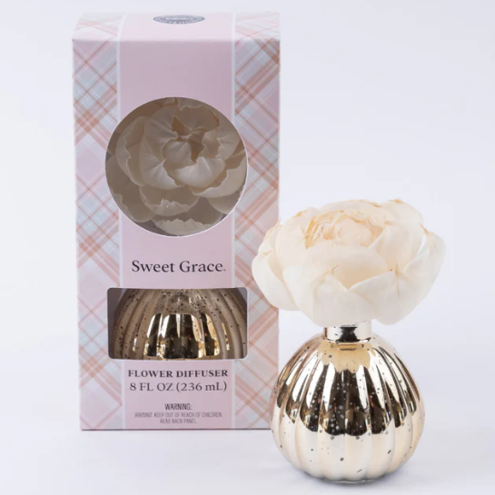 Gold flower diffuser with a white flower on top next to its packaging on a white background