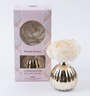 Gold flower diffuser with a white flower on top next to its packaging on a white background