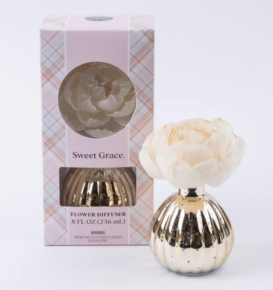 Gold flower diffuser with a white flower on top next to its packaging on a white background