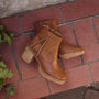 A pair of tan leather ankle boots with stacked wood-look block heels, shown from a top-down view on a red brick patio with green ivy leaves at the side.