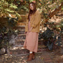 A woman stands on outdoor stone steps surrounded by lush green plants, wearing a tan utility jacket over a dusty pink maxi dress and brown ankle boots.