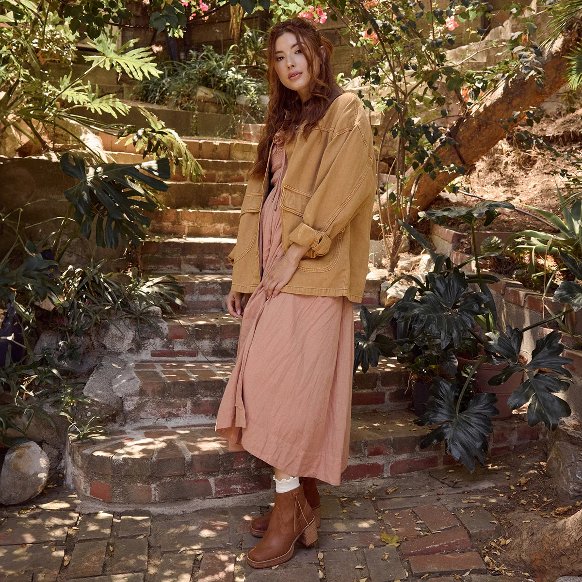 A woman stands on outdoor stone steps surrounded by lush green plants, wearing a tan utility jacket over a dusty pink maxi dress and brown ankle boots.