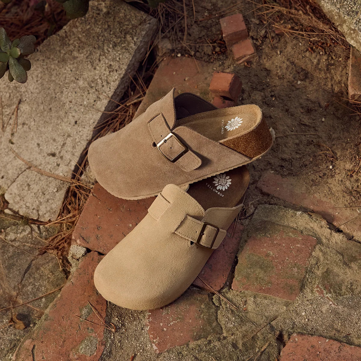 A pair of tan suede clogs with adjustable buckled straps, resting on rustic red bricks and concrete outdoors.