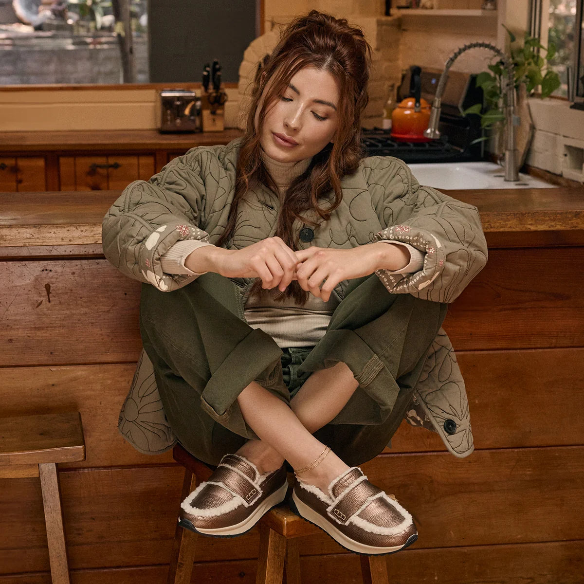 A woman sits on a wooden stool in a rustic kitchen setting, wearing a quilted olive green jacket, matching green pants, and metallic brown slip-on shoes with white faux-fur trim.