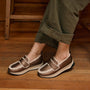 A person wearing brown slip-on loafers with white faux-fur trim and olive green cuffed pants, standing on a wooden floor.